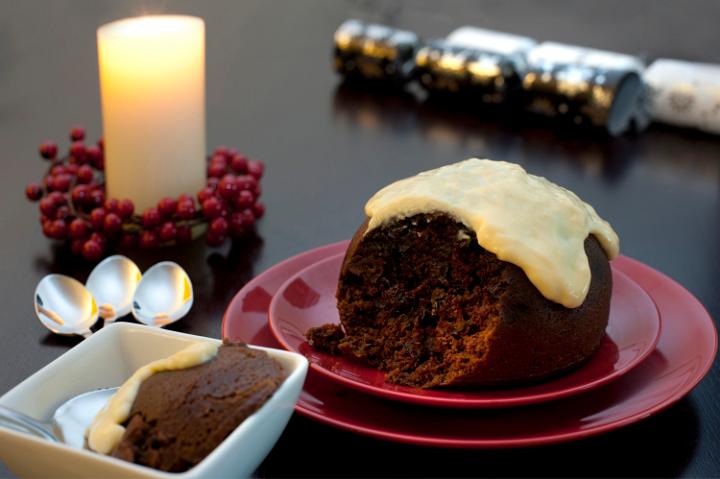Delicious fruity Christmas pudding with a brandy sauce topping and a serving in a bowl alongside on a festive Christmas table