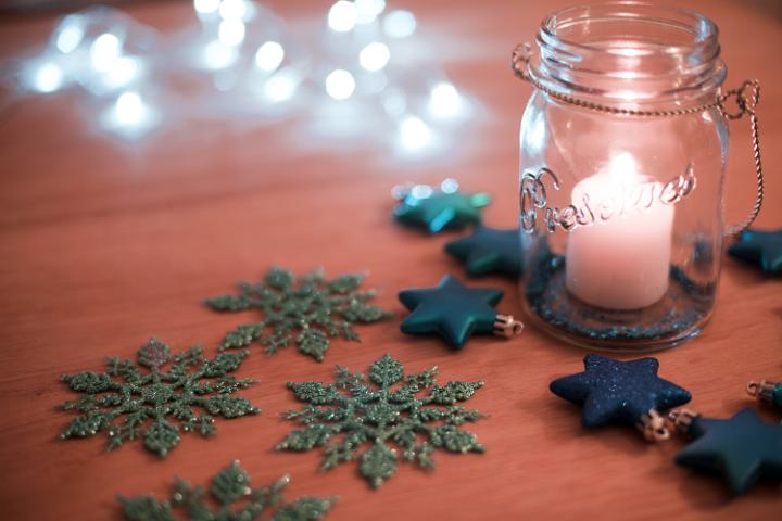 Burning Christmas candle in a glass jar casting a soft glow over surrounding snowflake and star decorations with a party light bokeh background