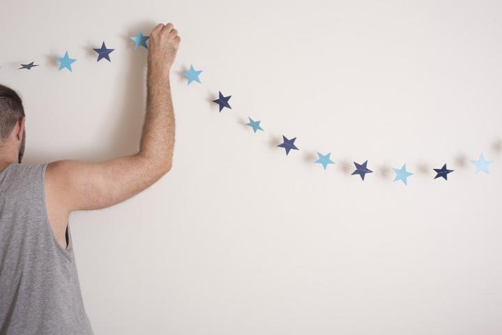 Man hanging simple Christmas bunting with a single string of blue paper stars on the wall of his house for a minimalist celebration