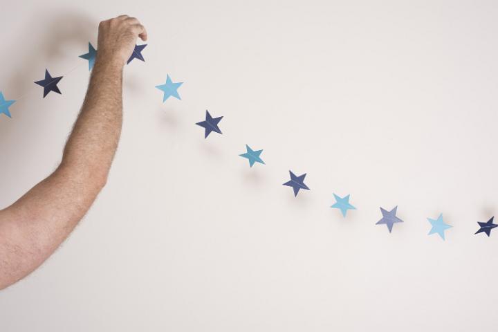 Man hanging decorative blue star bunting for Christmas at home against a white wall with copy space, close up of his bare arm