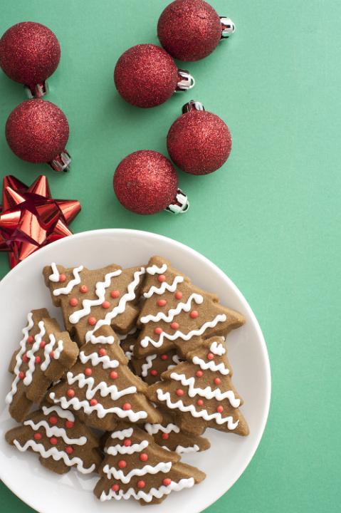 Plate of traditional homemade gingerbread Christmas cookies in the shaped of trees decorated with icing on a green background with baubles and bows, overhead view